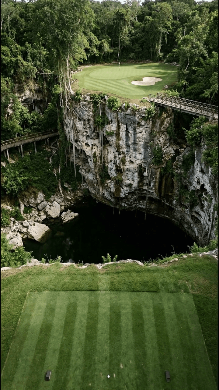 A short par 3 played from a striped tee across a vast circular limestone sinkhole to a green with one bunker on the far rim, sheer pale rock walls hung with vines falling hundreds of feet to still black water, jungle spilling over the edges and a timber walkway crossing on the right